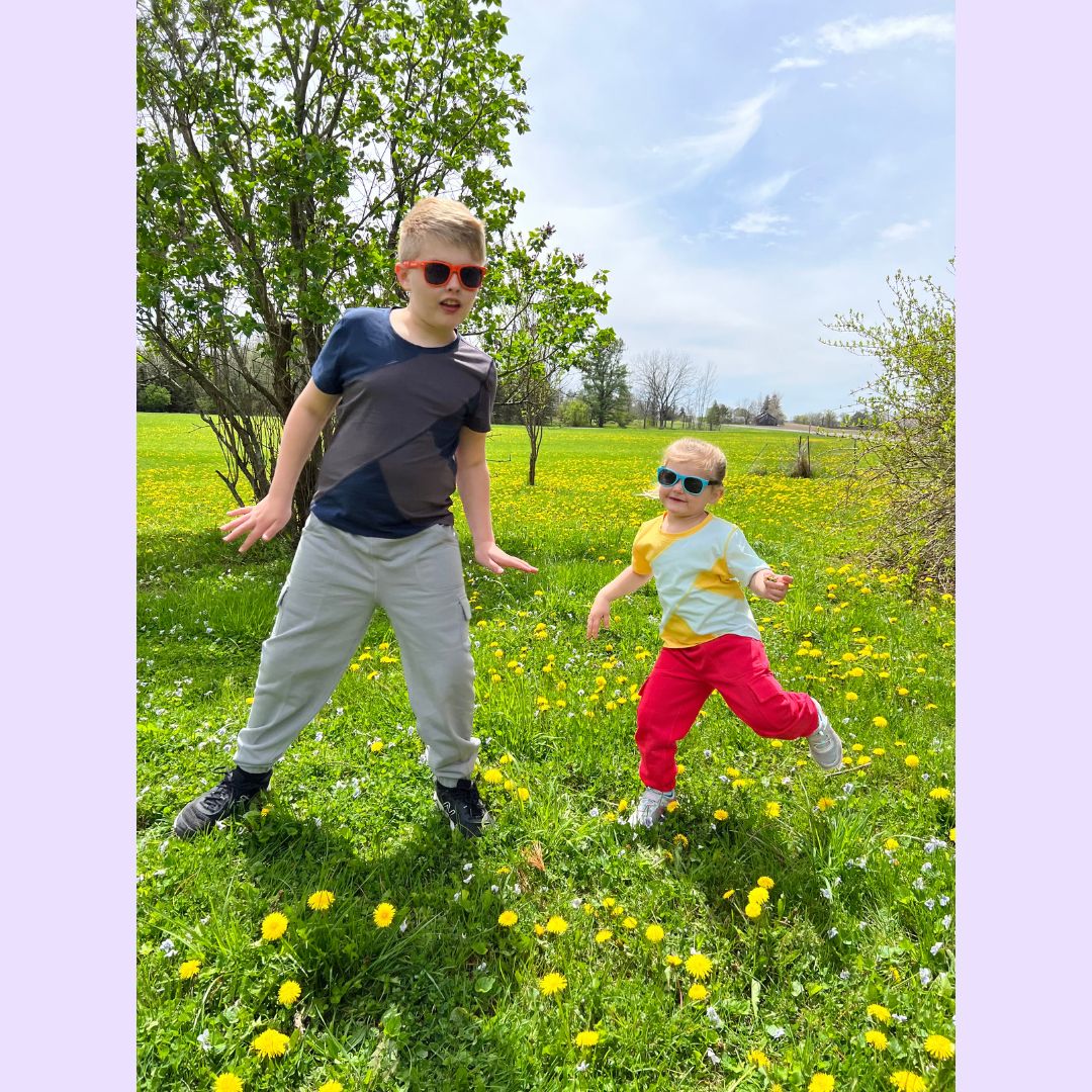 Two children playing in a grassy field with yellow flowers on a sunny day in colorful sensory clothing.