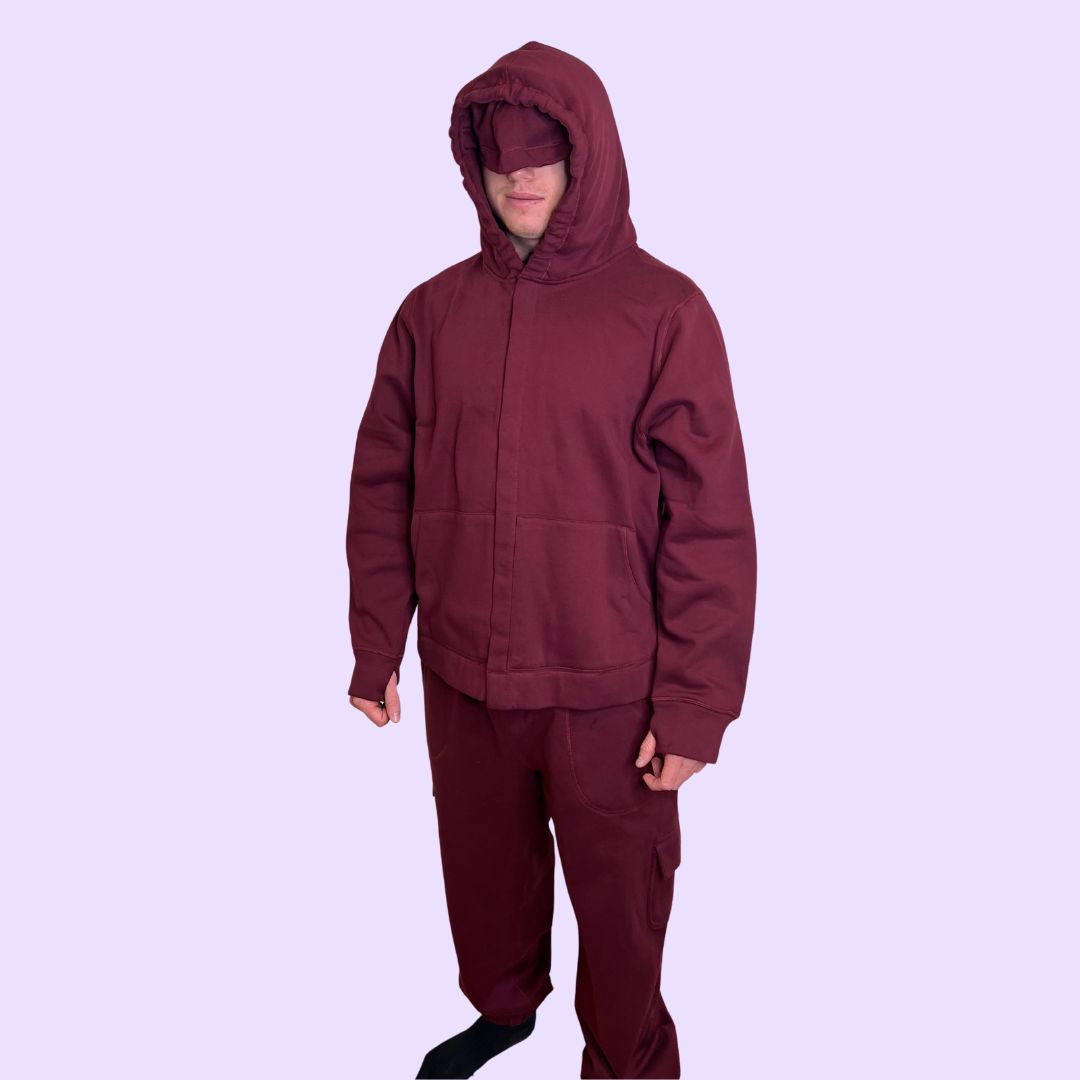 Adult man wearing a maroon sensory hoodie with eye mask down and pants on a light purple background.