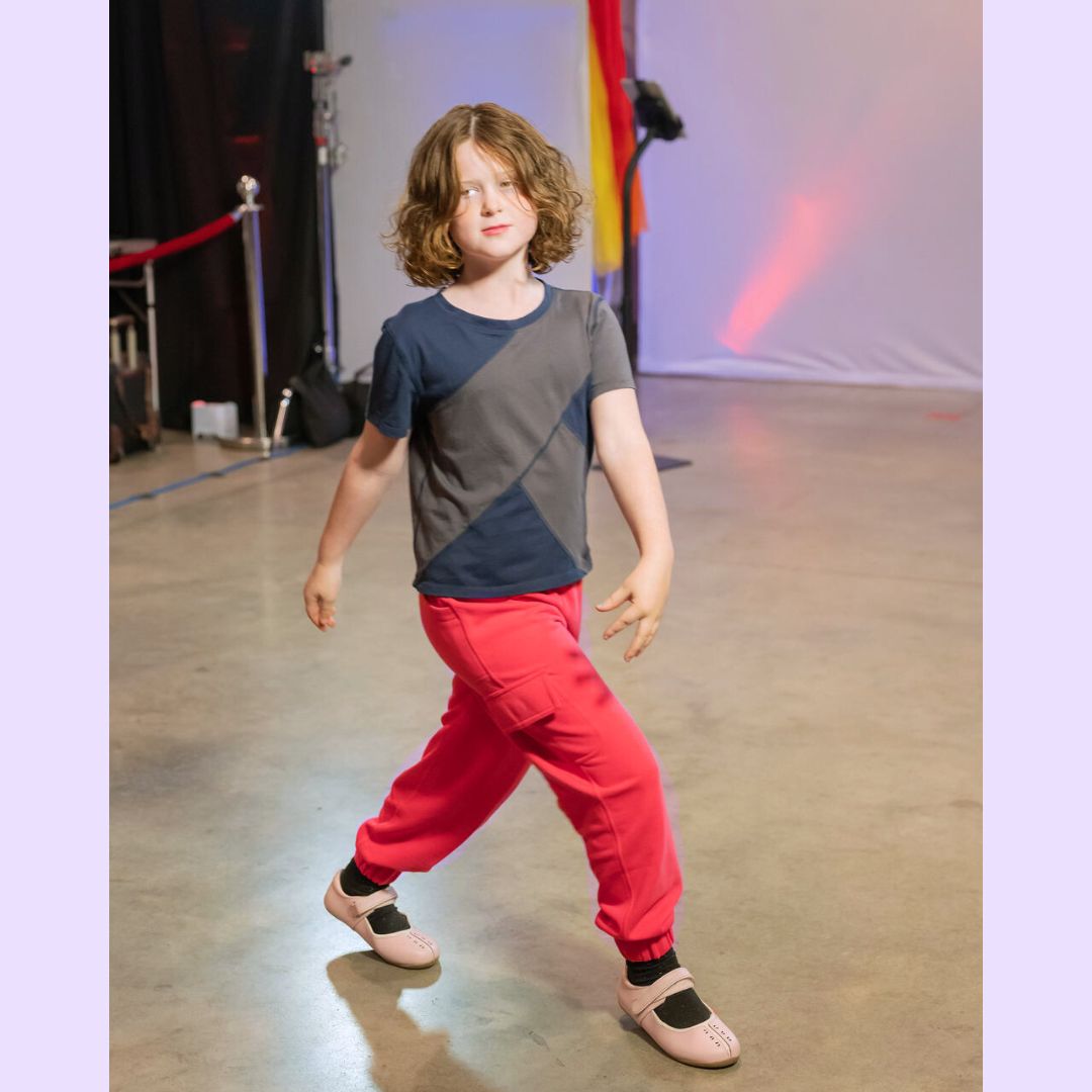 Child wearing a gray and navy shirt with coral pants on a stage modeling sensory clothing.