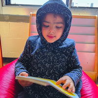Autistic child in a blue sensory sound reducing hoodie reading a book on a red beanbag chair.