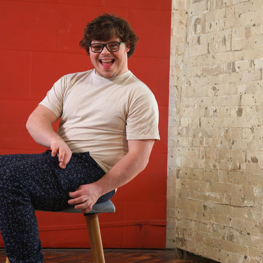 Man with down syndrome showing off fidget pocket of joggers while sitting in stool with red background.