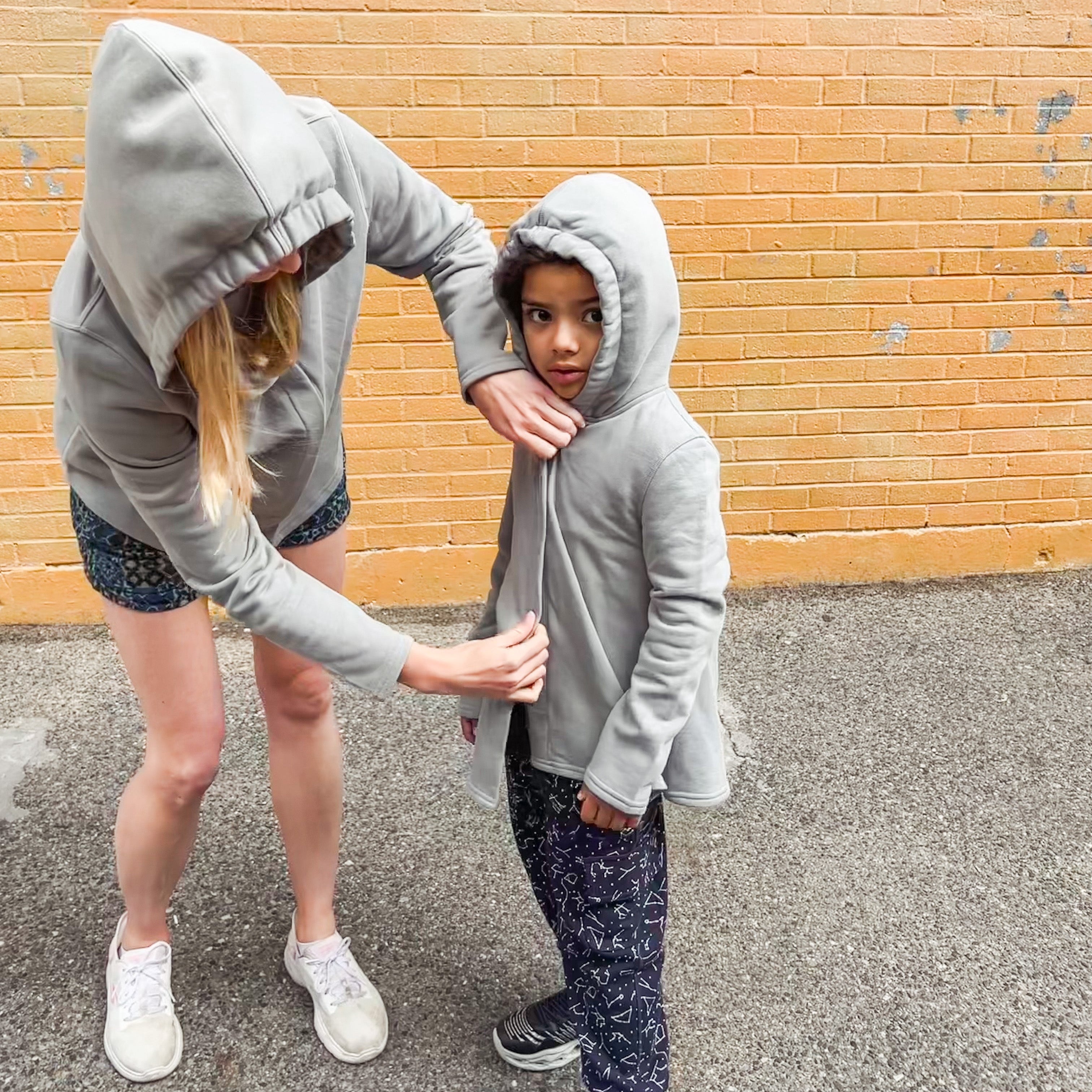 Teacher helping autistic student into their grey sound reducing sensory hoodies while wearing the same hoodie in adult sizes.