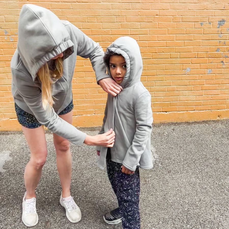 Teacher helping autistic student into their grey sound reducing sensory hoodies while wearing the same hoodie in adult sizes.